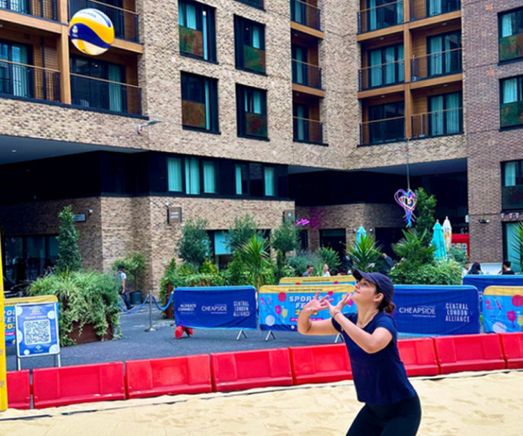 Woman practising a volleyball serve on a sand court set up outside residential buildings, part of a family fun day during Summer of Sport in the City.