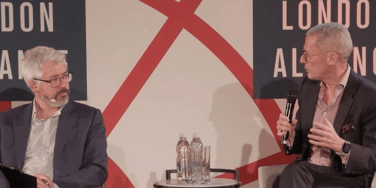 Two panelists seated in armchairs in conversation, one leaning forward speaking into a microphone with an animated hand gesture, in front of a red and white 'London Alliance' backdrop