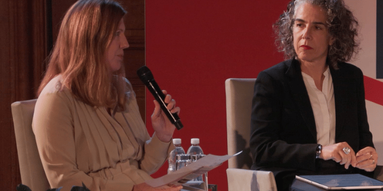 Two panelists seated close together in armchairs, one holding a microphone and papers while speaking, the other listening with hands clasped, against a plain backdrop