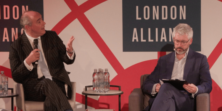 A speaker in a pinstripe suit gestures with his hand while speaking into a microphone, seated next to a panelist reviewing notes, in front of a red and white 'London Alliance' backdrop.